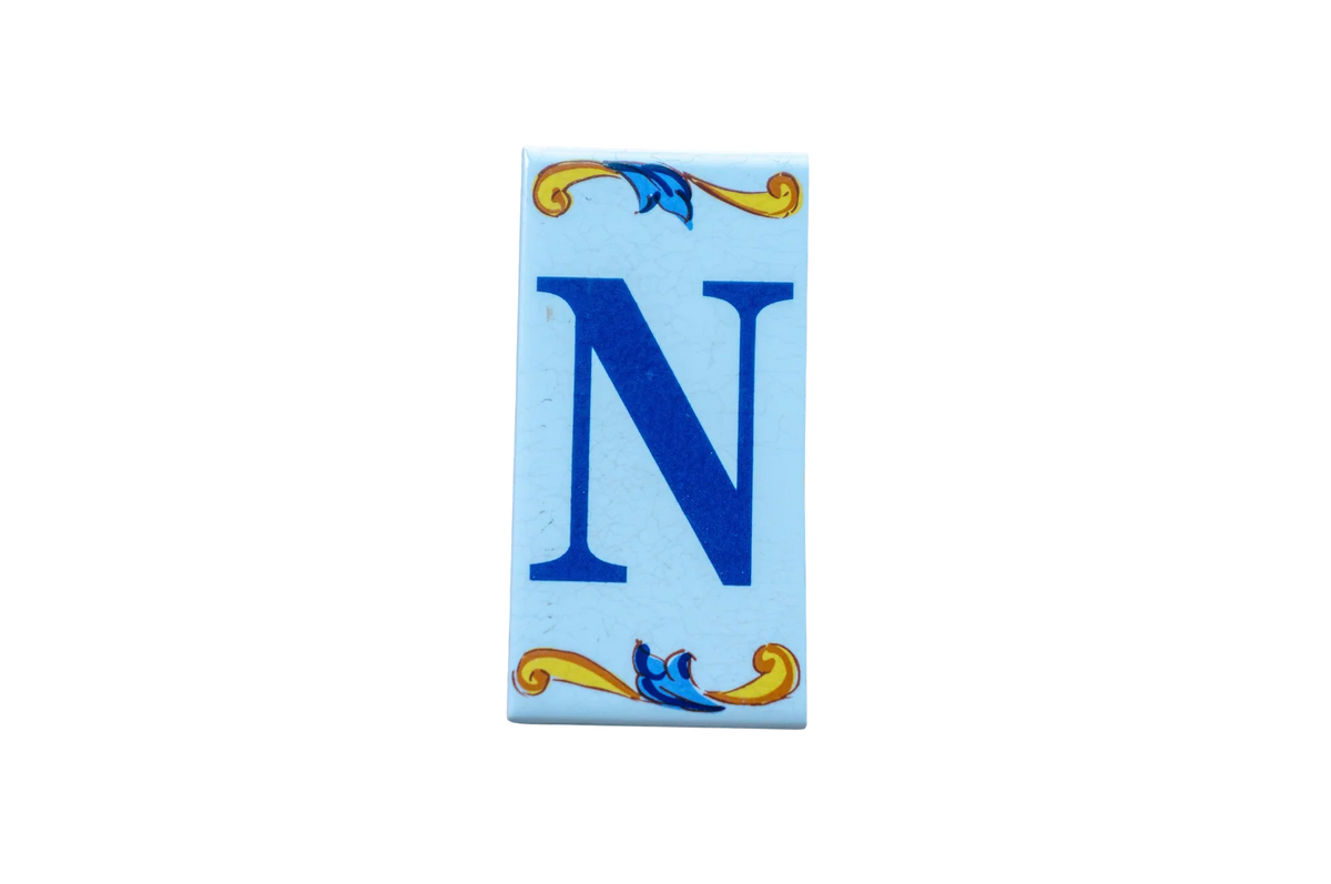 A rectangular Lisa Croquets Letters – Ceramic House Letter Tile featuring a blue capital letter N, decorated with blue and yellow scrollwork at the top and bottom.