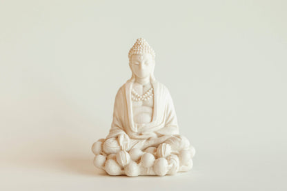 The Manulena Decorative & Aromatic Buddha Piece (White) sits cross-legged on a lotus base with detailed robes and beads, crafted from white ceramic, set against a plain light background.