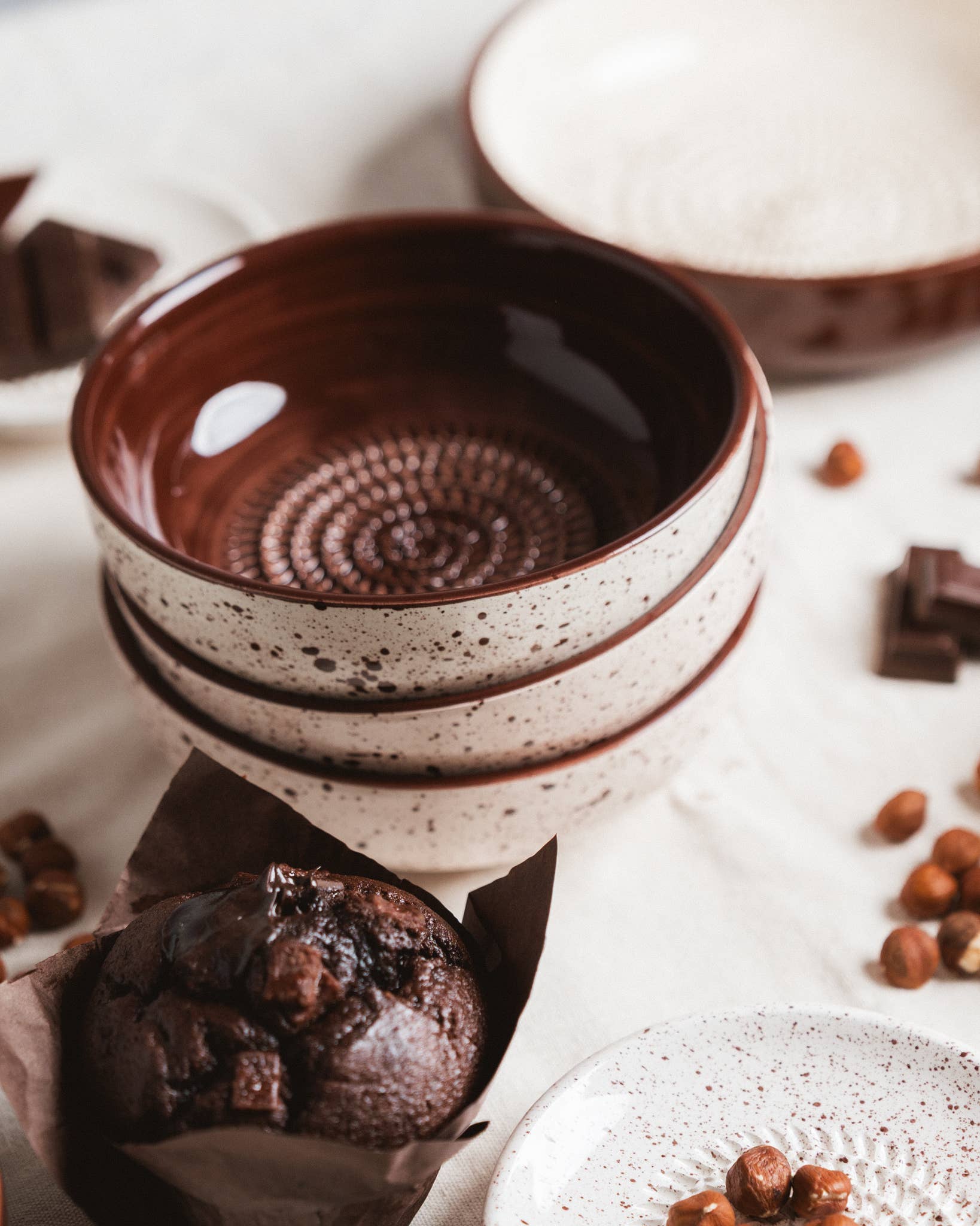 Three stacked Alfar Tierra Cocida Medium Pottery Grater Bowls in "Brownie" with brown interiors and speckled exteriors, arranged on a light surface with hazelnuts, chocolate pieces, and a chocolate muffin. Perfect for kitchen decor.