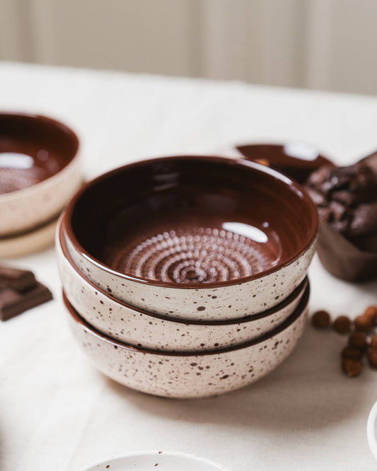 Three stacked Alfar Tierra Cocida Medium Pottery Grater Bowls in "Brownie," featuring a speckled beige exterior and glossy brown interior, rest on a white surface with other dishes and food items partially visible nearby.