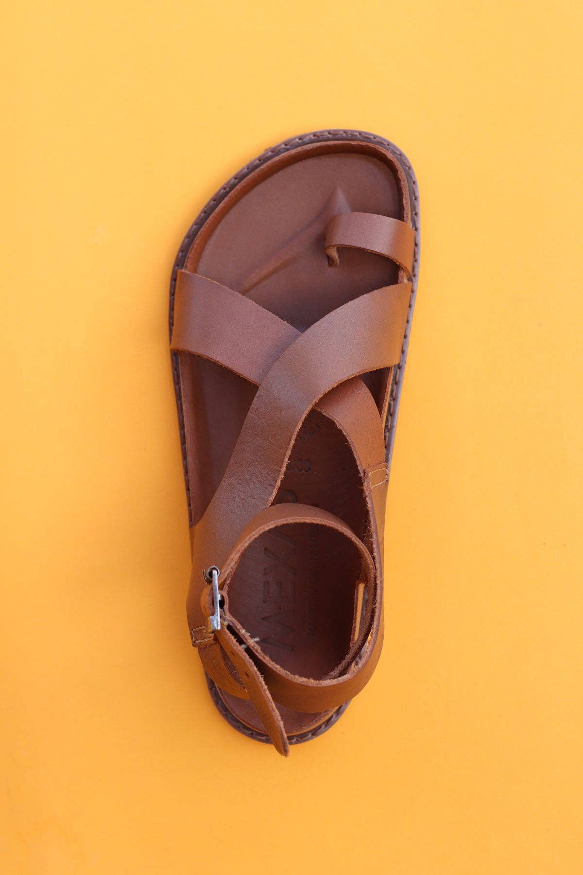 A pair of brown leather Tampico sandals by Mexas EU, featuring criss-cross straps and a buckle, is shown from above on a solid yellow background.
