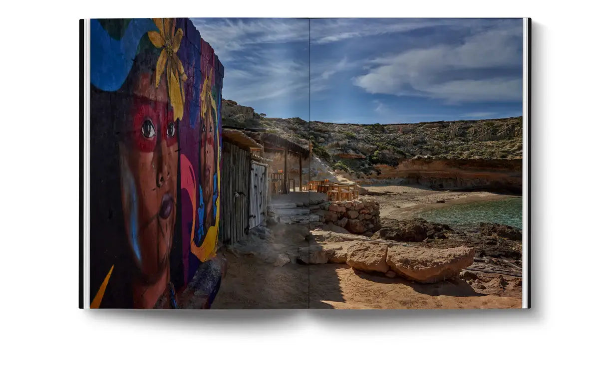 A vibrant mural beside rustic huts on a rocky Ibiza beach under clear blue sky and calm water—a stunning scene featured in "The Coolest Ibiza" by Loft Publications, perfect for any stylish coffee table.