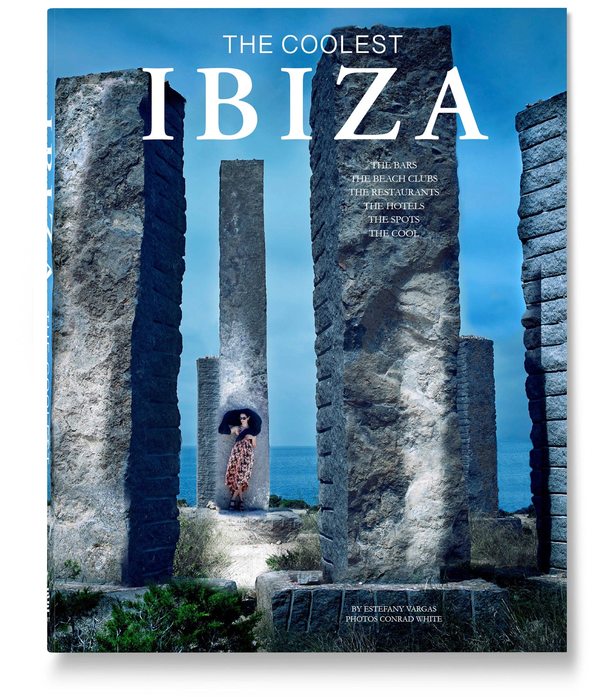 A woman in a patterned dress stands among stone pillars under a blue sky on the cover of The Coolest Ibiza by Loft Publications, a photography book showcasing Ibiza’s top beach clubs, restaurants, hotels, and attractions.
